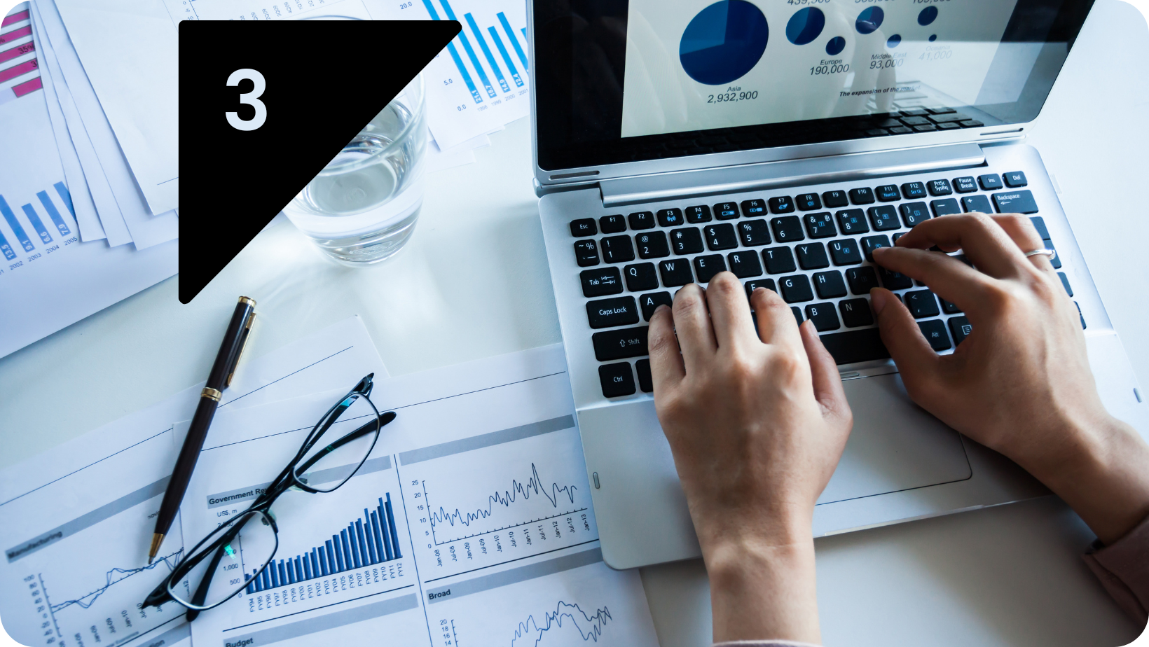 A selection of reports on a desk and a laptop with a pair of glasses and a glass of water set to one side